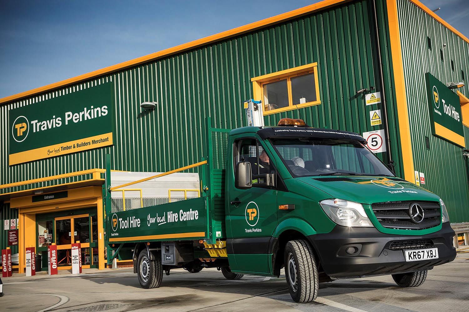A green Travis Perkins Tool Hire flatbed truck is parked in front of a matching green Travis Perkins building.