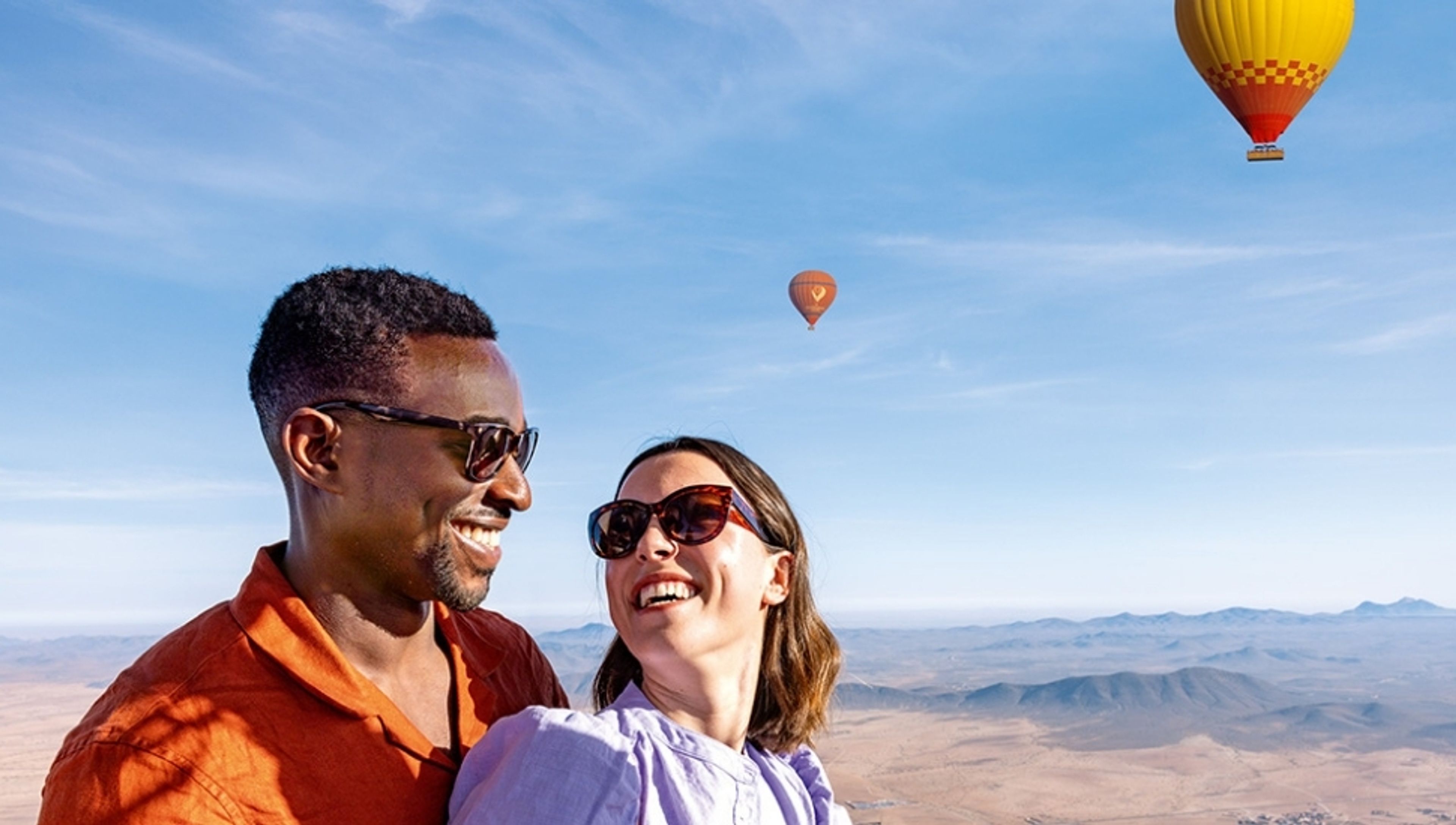 A smiling couple in a hot air balloon, overlooking a desert landscape with other balloons in the sky.