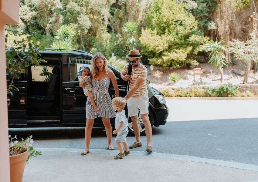 A family with two young children stands next to an open black minivan on a sunny day.
