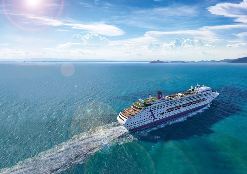 A white and purple cruise ship sails on turquoise water, leaving a wake, under a bright, sunny sky with distant islands.