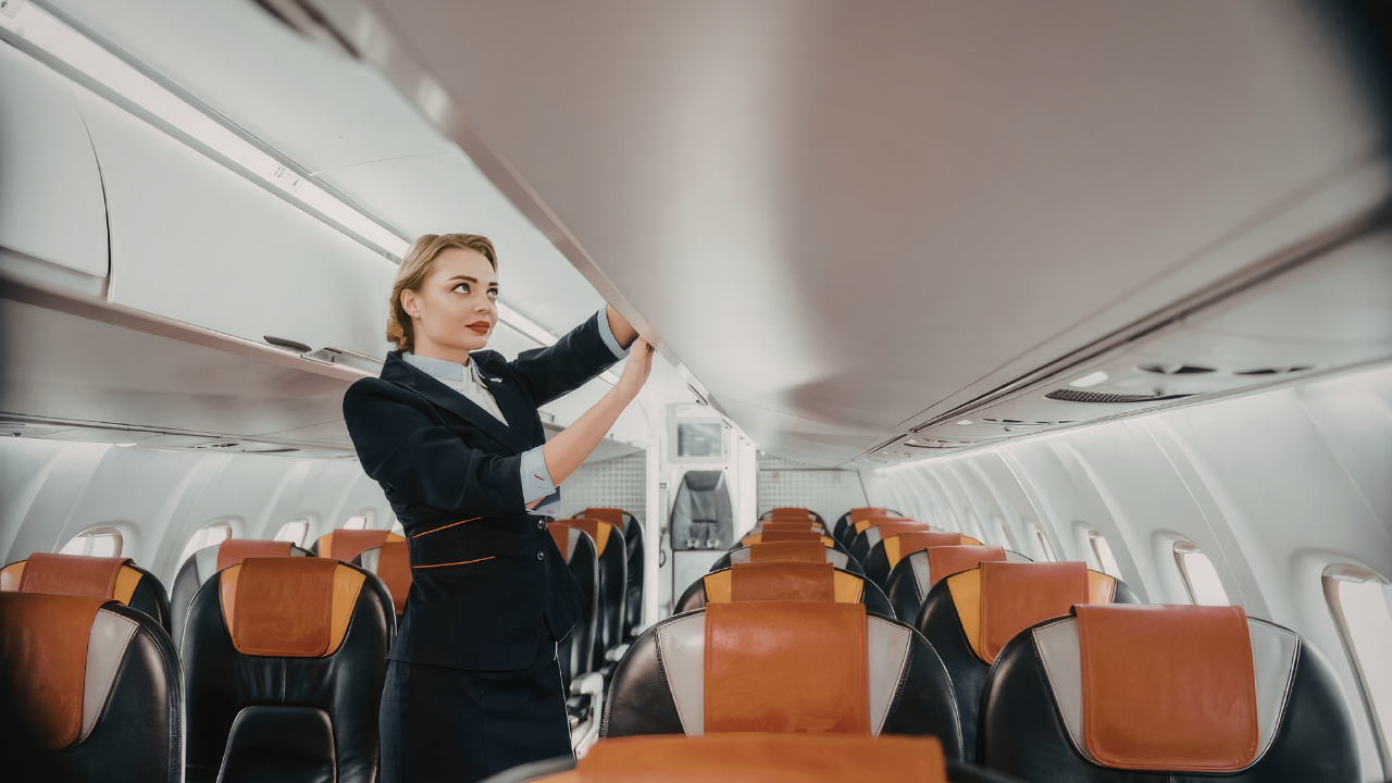 A flight attendant in uniform closing an overhead bin in an airplane cabin with empty seats.
