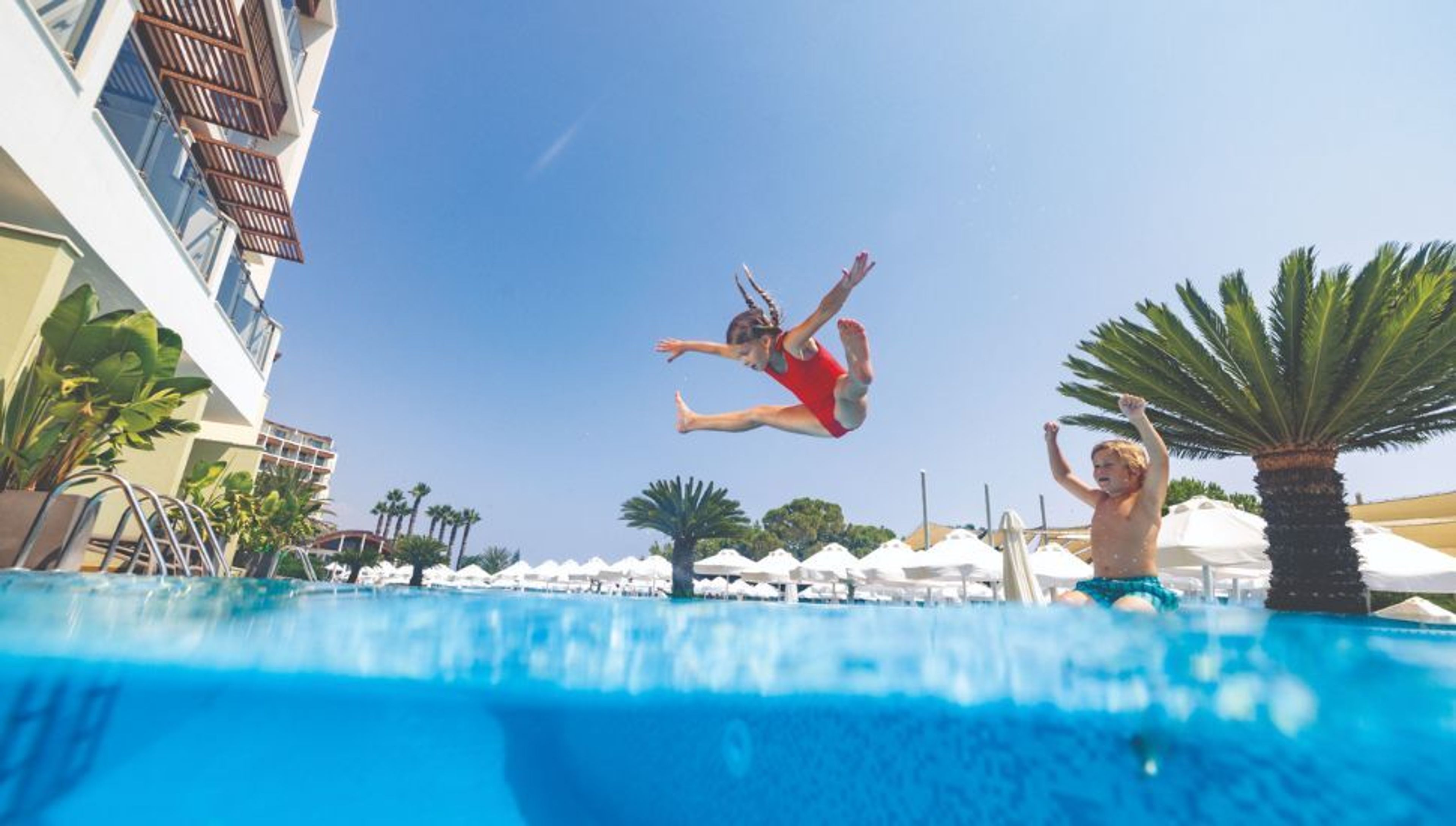 A girl leaps into a swimming pool as a boy cheers, from a half-above, half-below water perspective with a resort in the background.