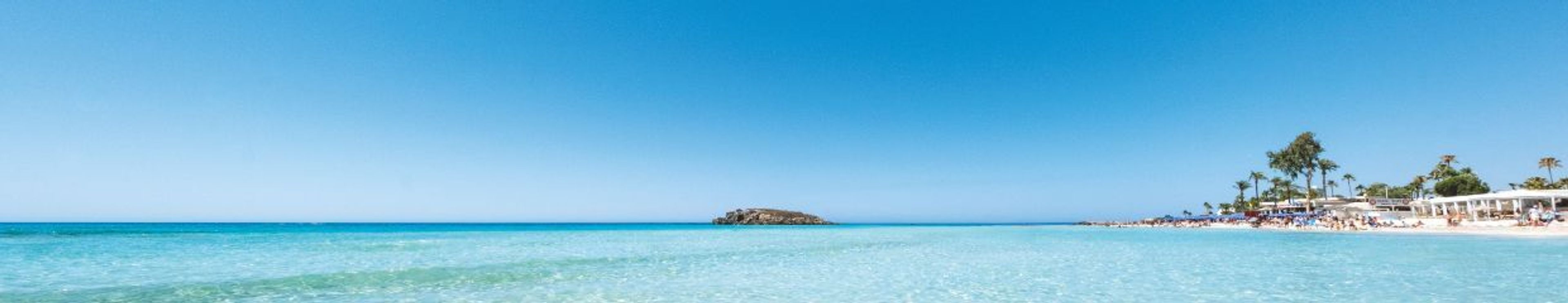 A panoramic beach scene with clear turquoise water, a rocky outcrop, and a sandy shore lined with a resort and palm trees under a bright blue sky.