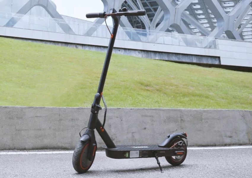 A black electric scooter with orange accents is parked on pavement with a grassy slope and modern building in the background.