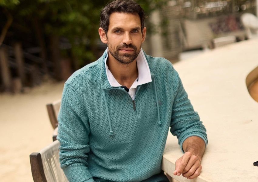 A man with a beard wearing a teal knitted quarter-zip sweater sits at an outdoor table.