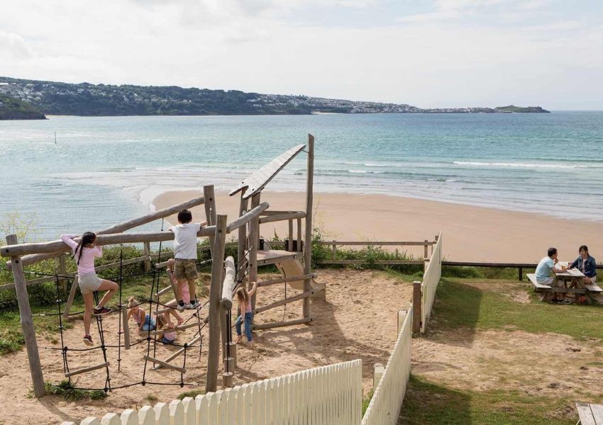 Children playing on a wooden playground overlooking a sandy beach and ocean, with adults at a picnic table.