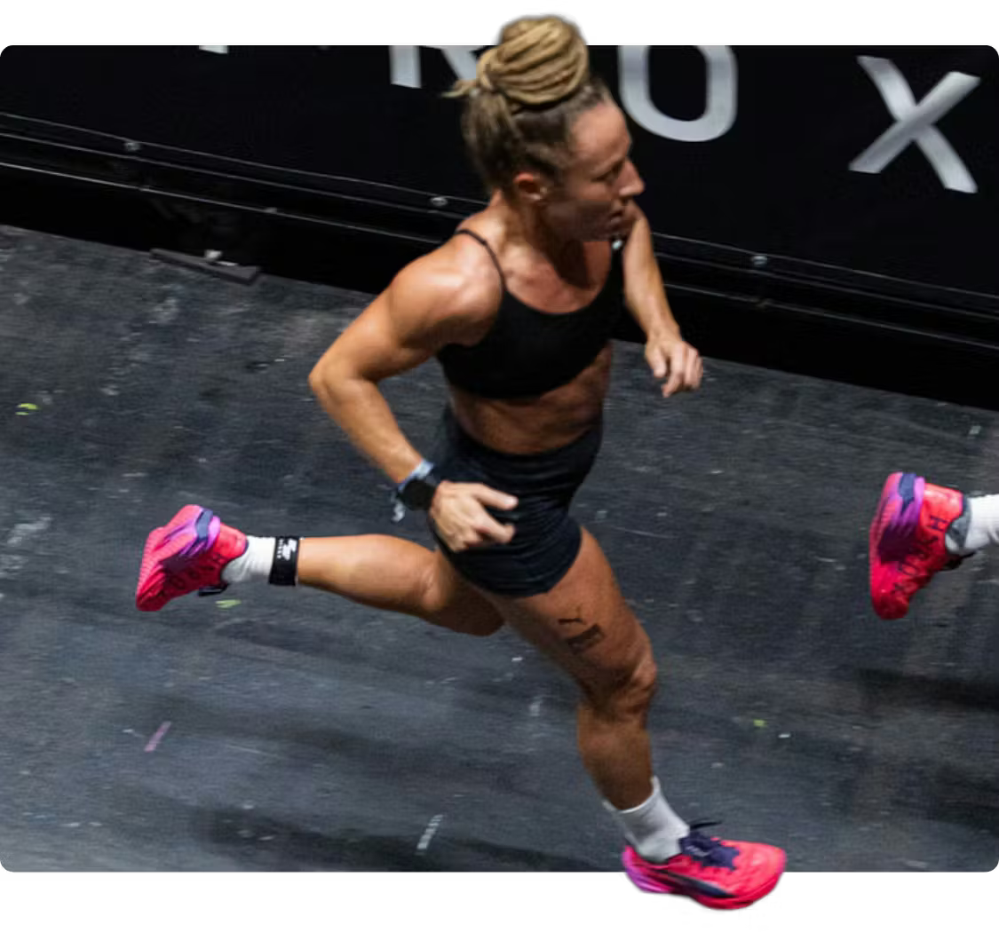 A woman running in a black sports bra, black shorts, and bright pink shoes.