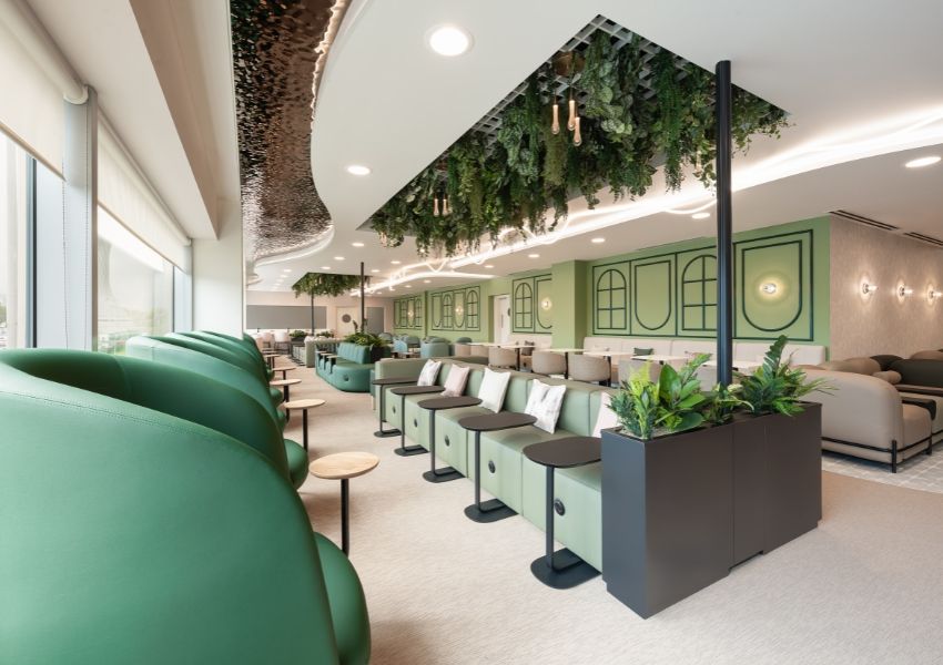 A bright, modern lounge featuring rows of green seating, decorative mint green walls, and ceiling cutouts filled with lush hanging plants.