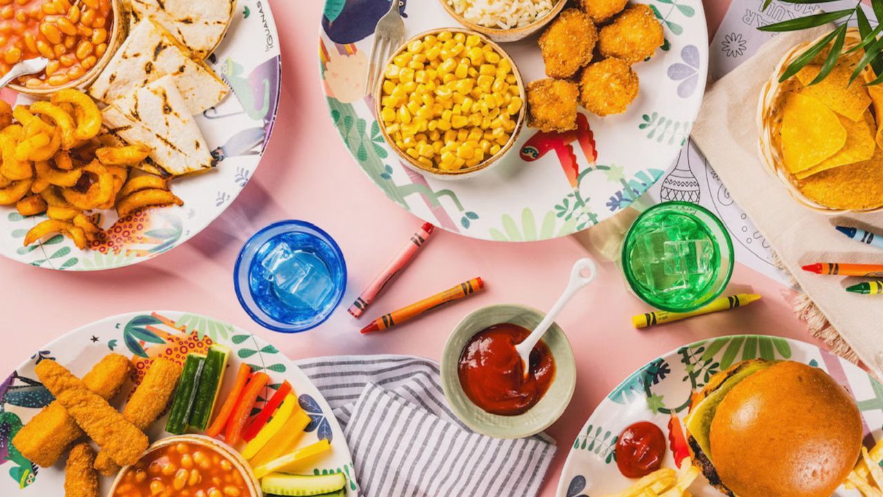 A pink table with a spread of kid-friendly meals, including burgers, fries, nuggets, vegetables, corn, drinks, crayons, and coloring pages.