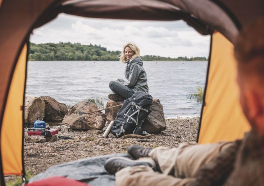 View from inside a tent of a smiling woman sitting on rocks by a lake, with a backpack and stove nearby.