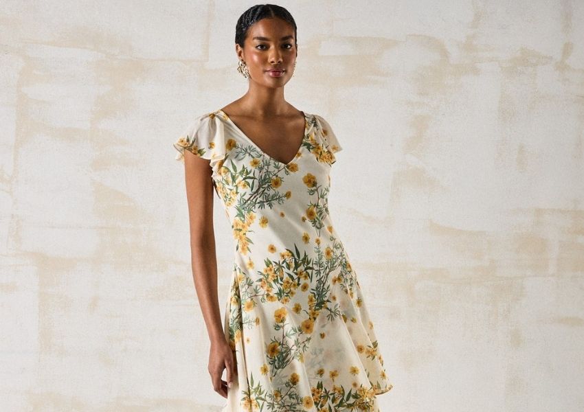 A dark-skinned woman in a white V-neck dress with yellow floral print and flutter sleeves.