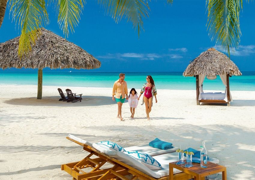 Family walking on a tropical white sand beach with turquoise water, lounge chairs, and straw umbrellas.