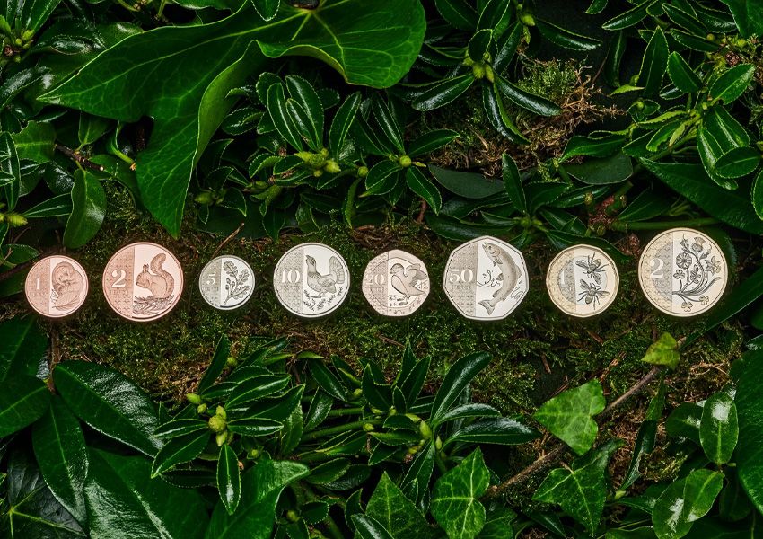 A row of eight coins featuring various animals and plants, laid on moss and green leaves.
