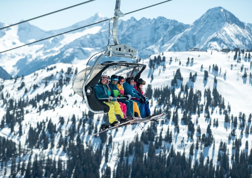 Skiers in a ski lift above a snowy mountain landscape with pine trees.
