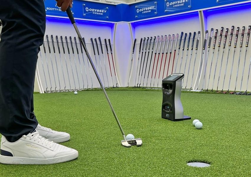 A person putts a golf ball on an indoor green with a putting analysis device and golf clubs displayed in the background.