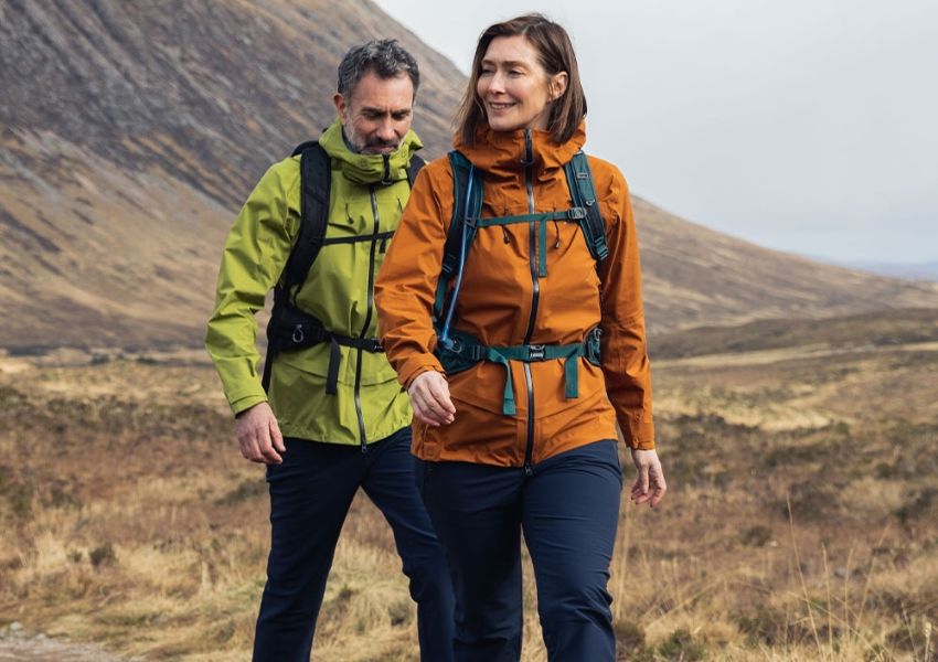 A smiling woman in an orange jacket and a man in a green jacket hike on a trail in a dry, mountainous landscape.