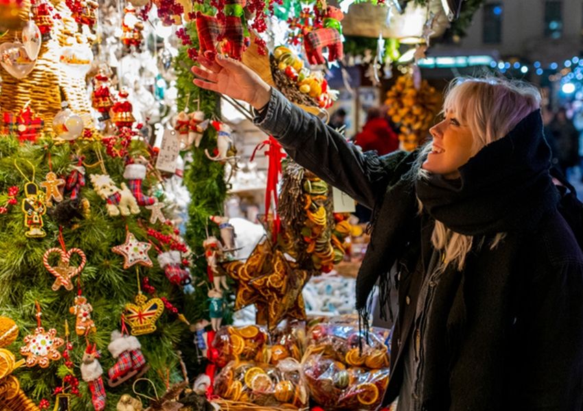 Smiling woman reaching for an ornament at a Christmas market stall with festive decorations and dried orange slices.