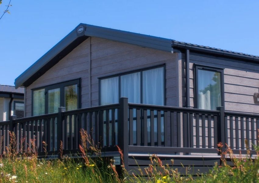 A modern gray lodge with a dark deck, seen from a low angle with wildflowers in the foreground under a blue sky.