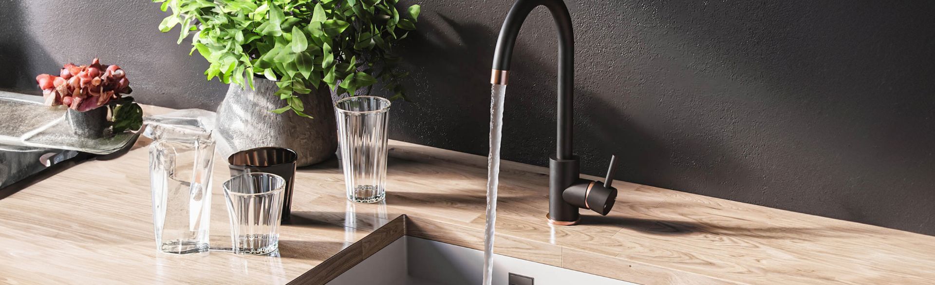 Water streams from a modern black and copper kitchen faucet into a sink, with glasses and plants on a wooden counter.