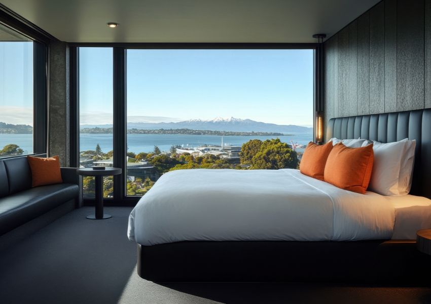 A modern hotel room with a bed and sofa, featuring a large window view of a bay, town, and distant snow-capped mountains.