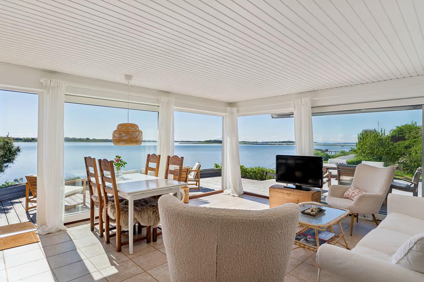Bright sunroom with dining and living areas, overlooking a tranquil lake through large windows.