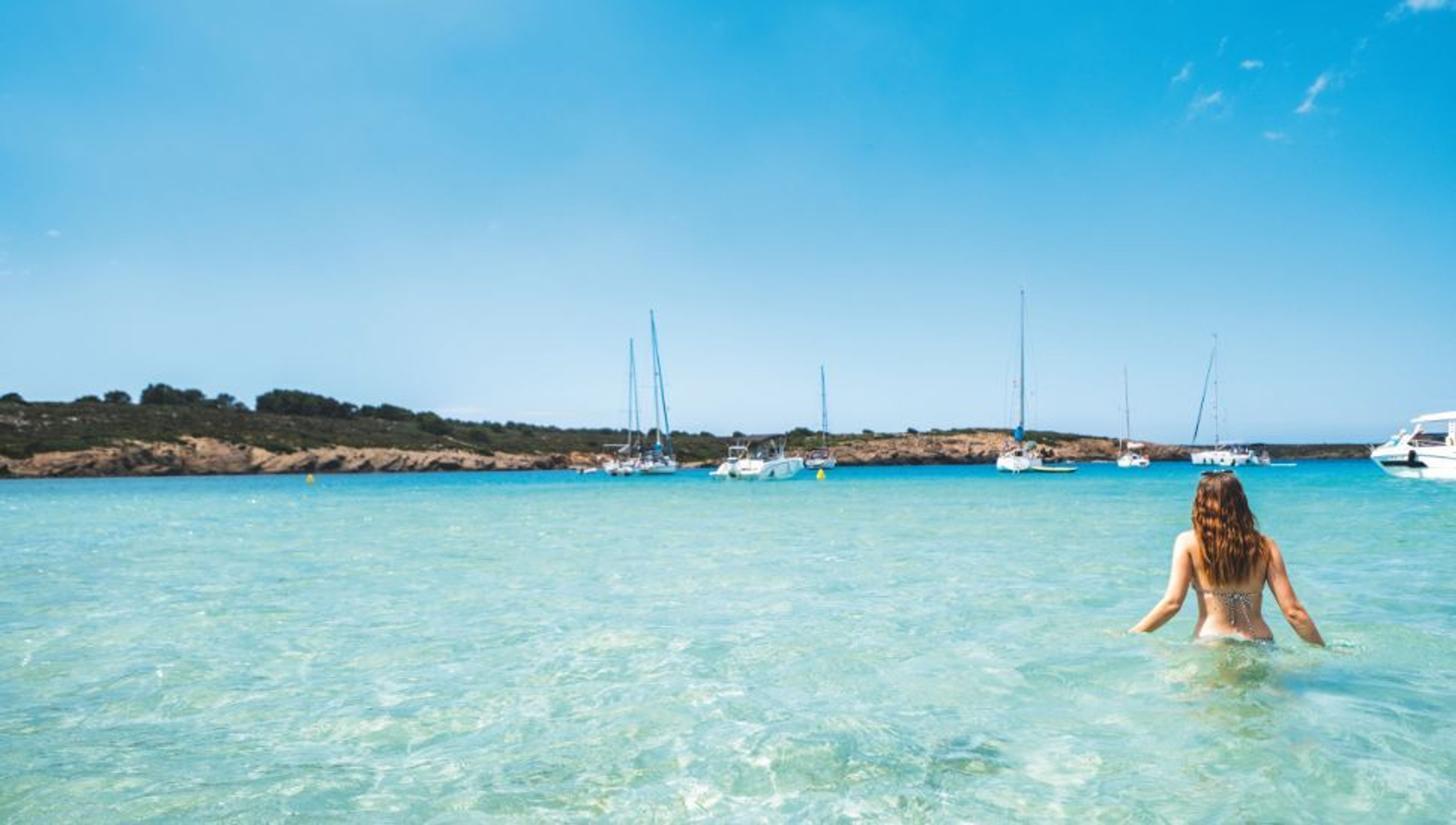 Woman wading in clear turquoise water with sailboats and a green coastline in the background.