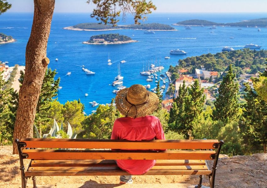 A person in a straw hat sits on a bench overlooking a bay with boats, a coastal town, and green islands.