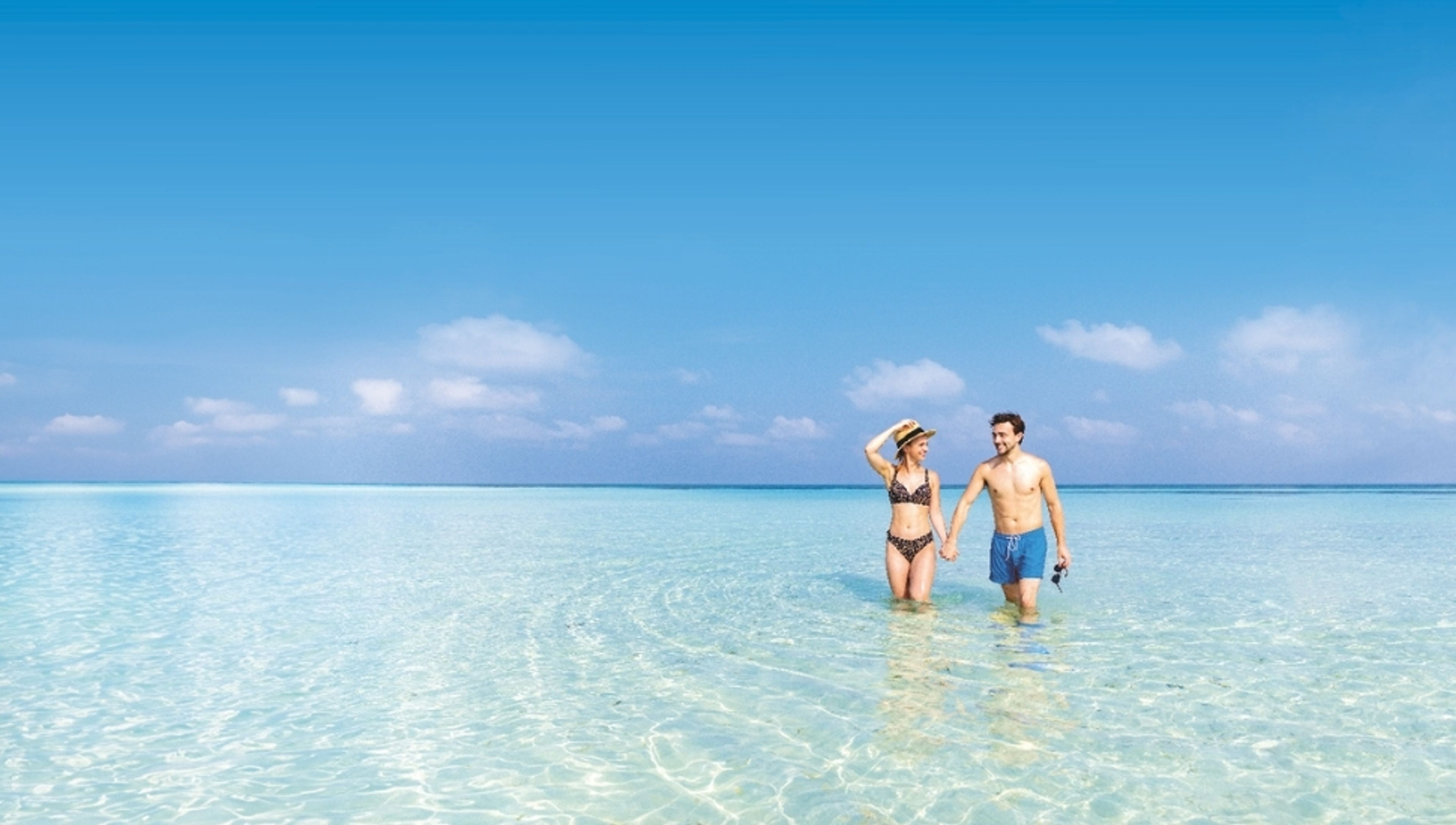 A couple holding hands and walking in clear, shallow turquoise water under a blue sky.