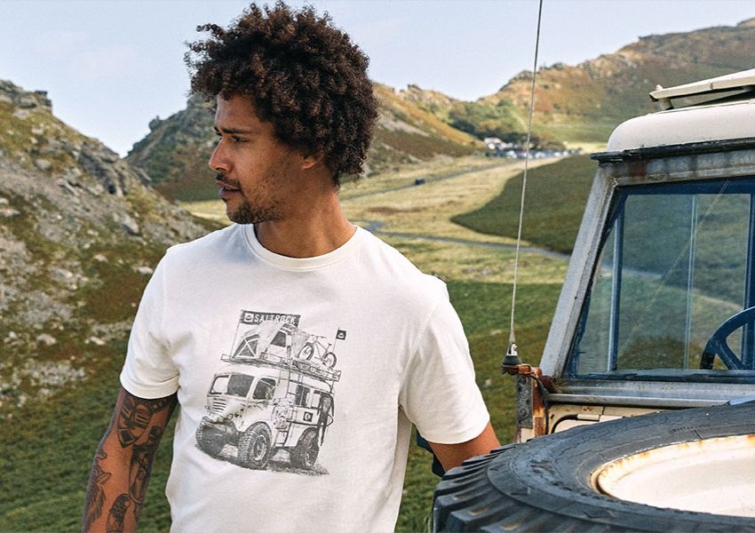A man with curly hair and a beard wears a white t-shirt with a graphic of an overland truck, standing by an old 4x4 in a mountainous setting.