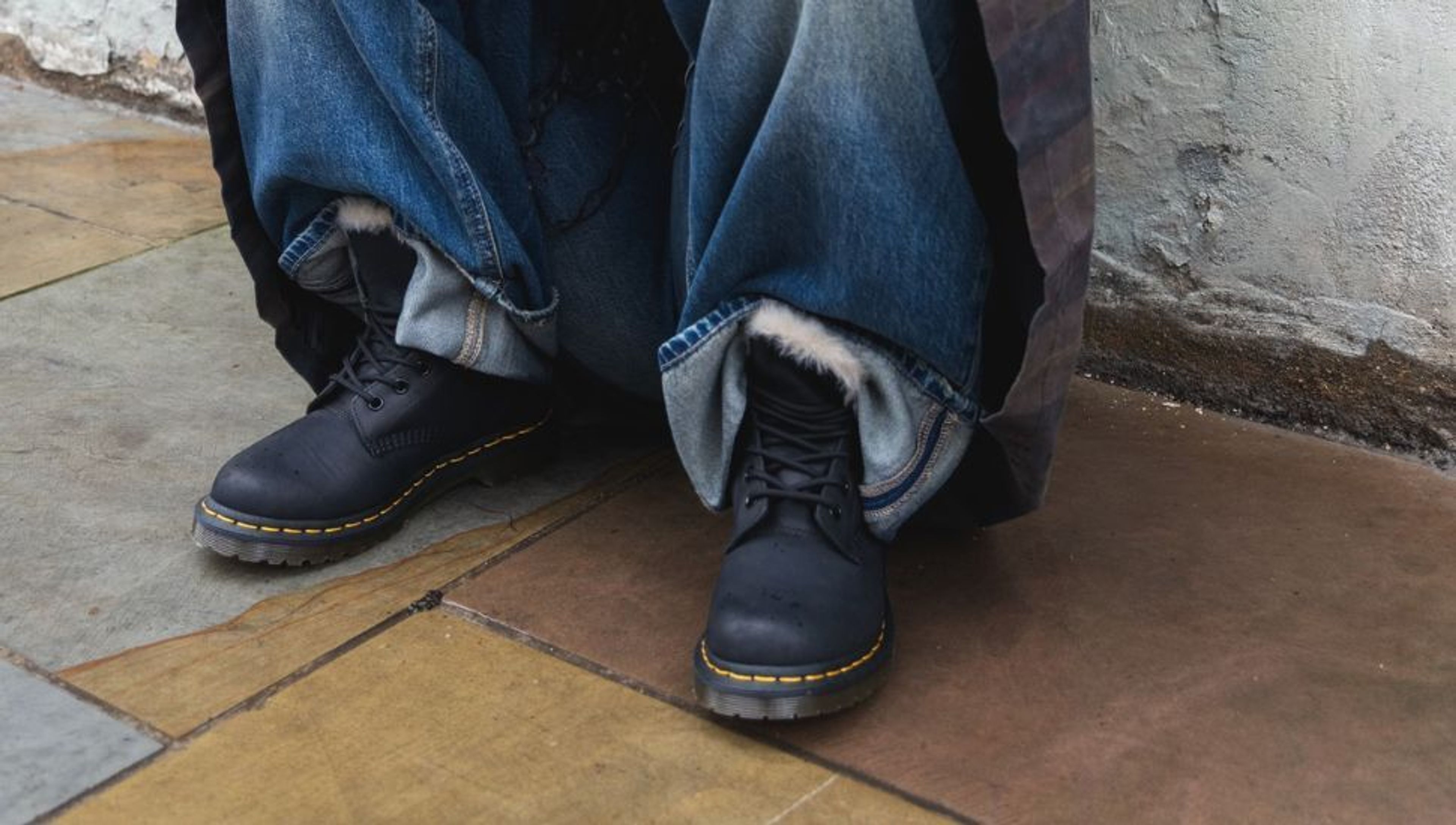 A person's feet wearing dark boots with yellow stitching and wide, fur-lined denim jeans on a tiled floor.
