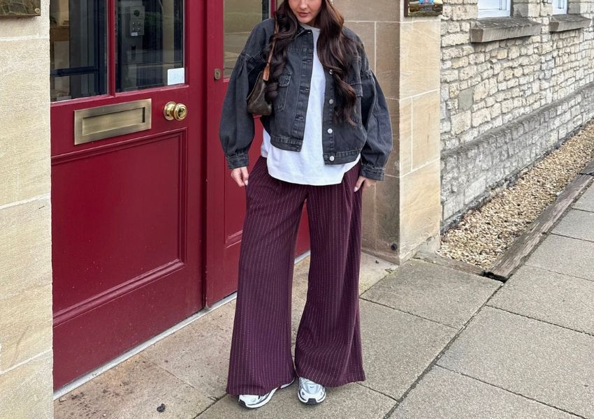 A person in a black denim jacket, white t-shirt, and maroon wide-leg pleated pants stands outside a red door.