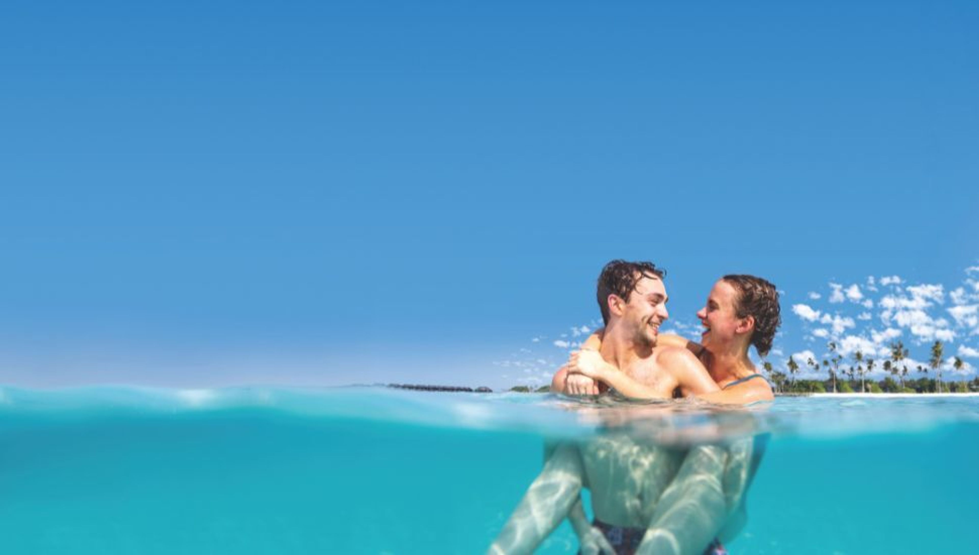 A smiling couple embraces in clear blue ocean water, with a tropical island visible in the background.