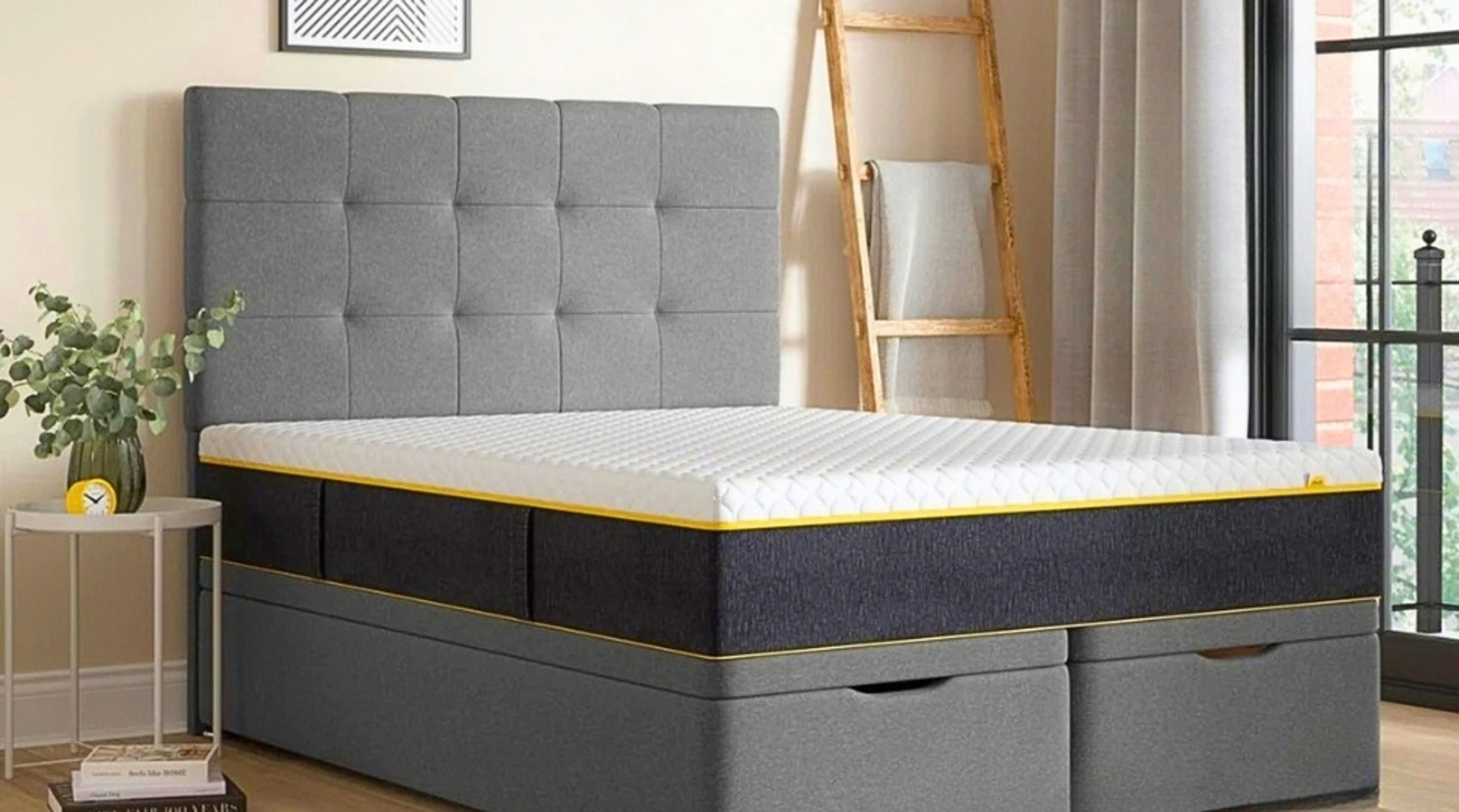 A grey upholstered bed with storage drawers and a mattress featuring white, dark grey, and yellow.
