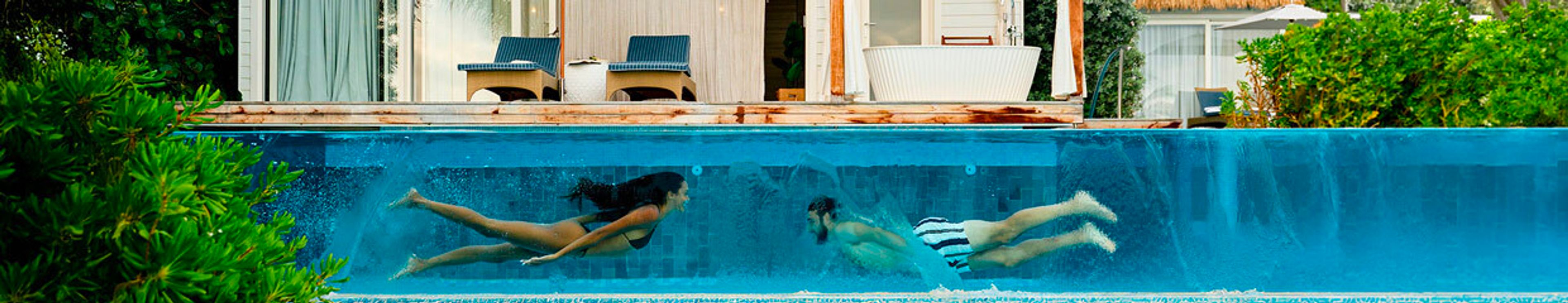 Two people swim underwater in a transparent-sided pool, with a modern house and outdoor bathtub visible above.
