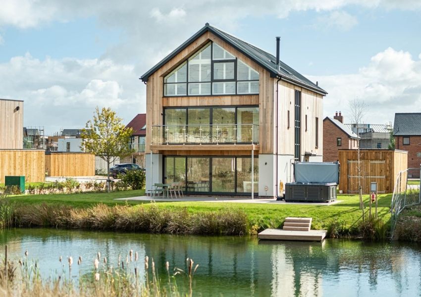 A modern wood-clad house with large windows and a balcony overlooks a pond, featuring a hot tub and a small dock on the lawn.