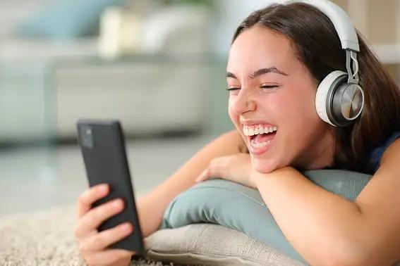 Young woman wearing white headphones, laughing at her smartphone.