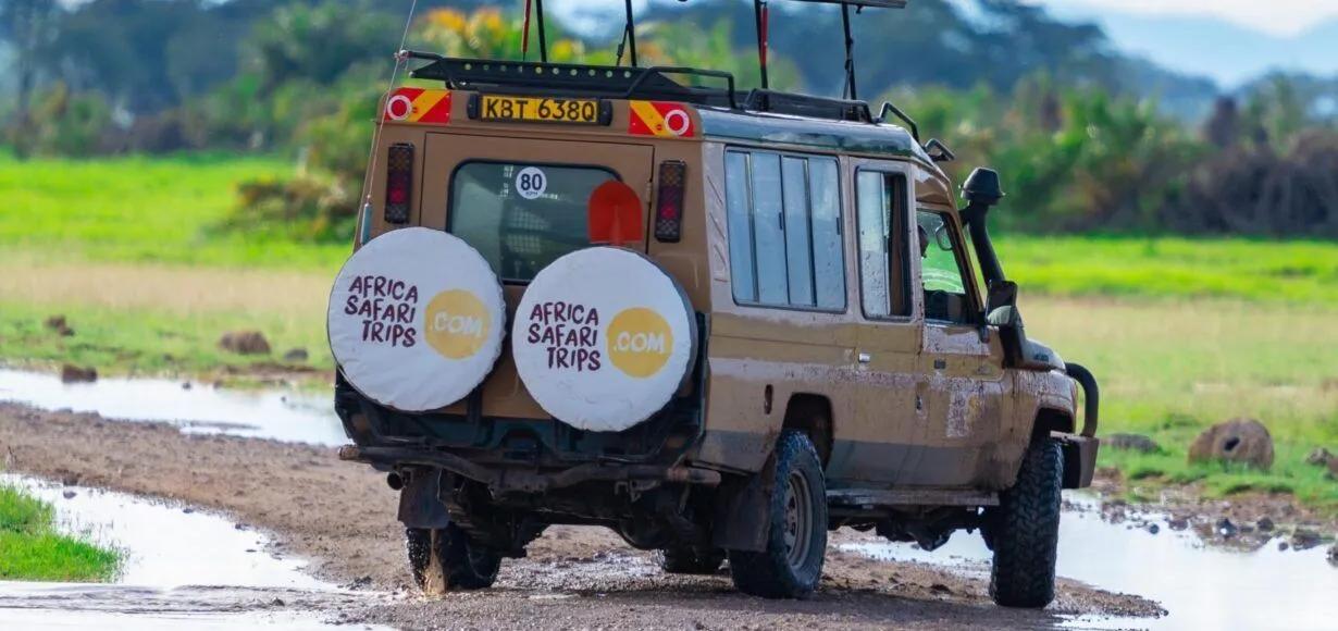 A dirty tan safari vehicle with "Africa Safari Trips" branding drives on a muddy road through a green landscape.