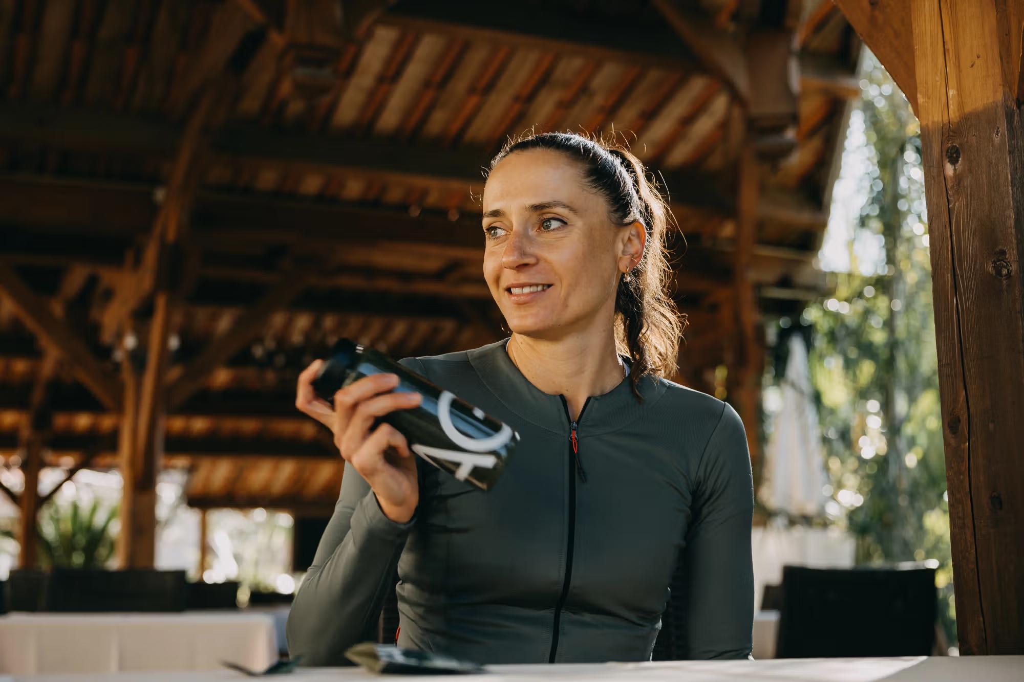 Smiling woman in athletic wear holding a black water bottle under a wooden pavilion.