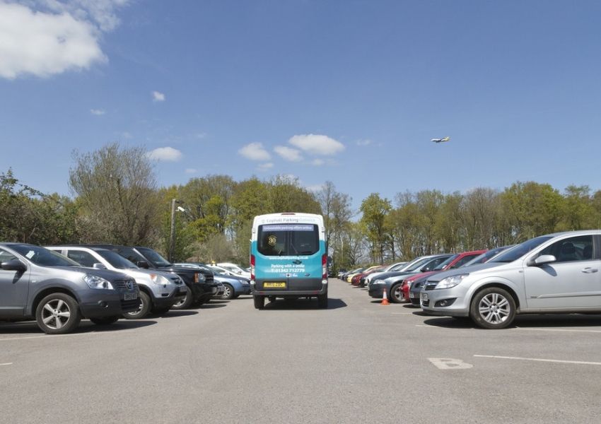 A parking lot full of cars with a shuttle van in the center and an airplane flying under a blue sky.