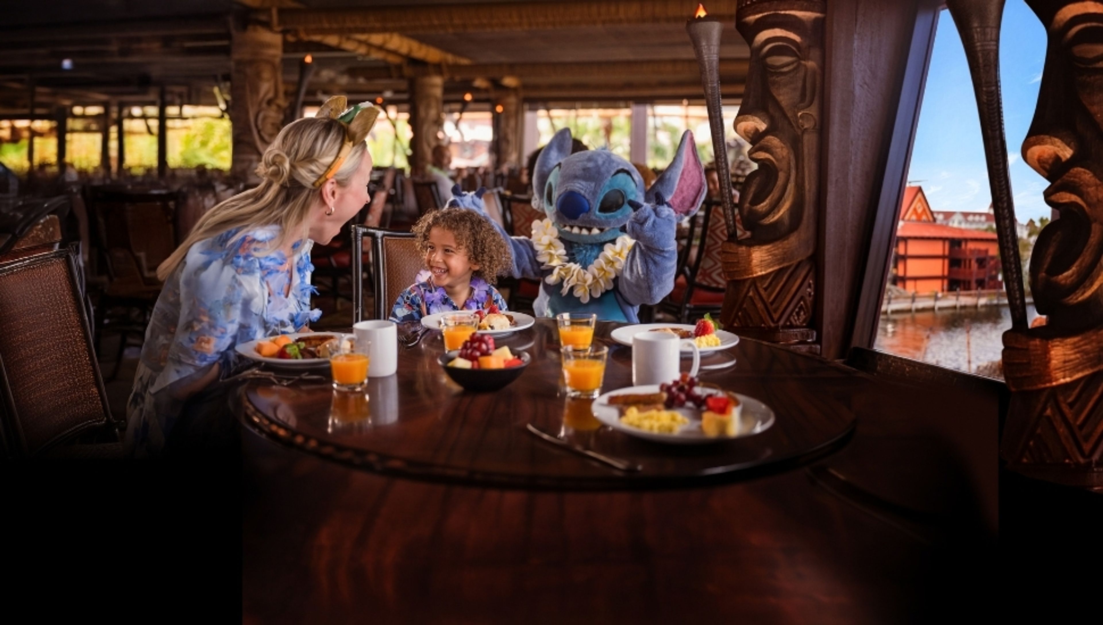 A woman and child laugh with Stitch at a character breakfast in a Polynesian-themed restaurant with a water view.