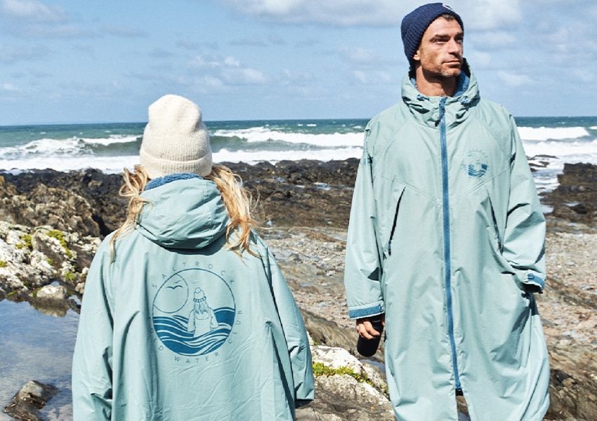 Two people in full-length teal robes and beanies on a rocky beach by the ocean.
