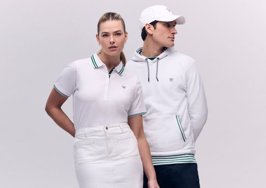 A woman in a white polo shirt with green trim and a white skirt stands next to a man in a white hoodie with green trim, dark pants, and a white baseball cap.