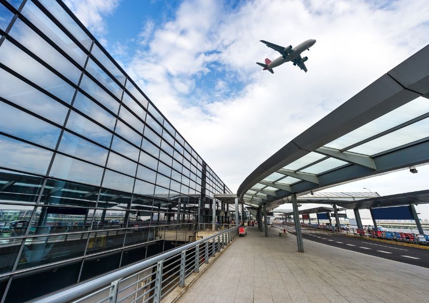An airplane ascends over a modern airport terminal building with a glass facade and a covered outdoor walkway.
