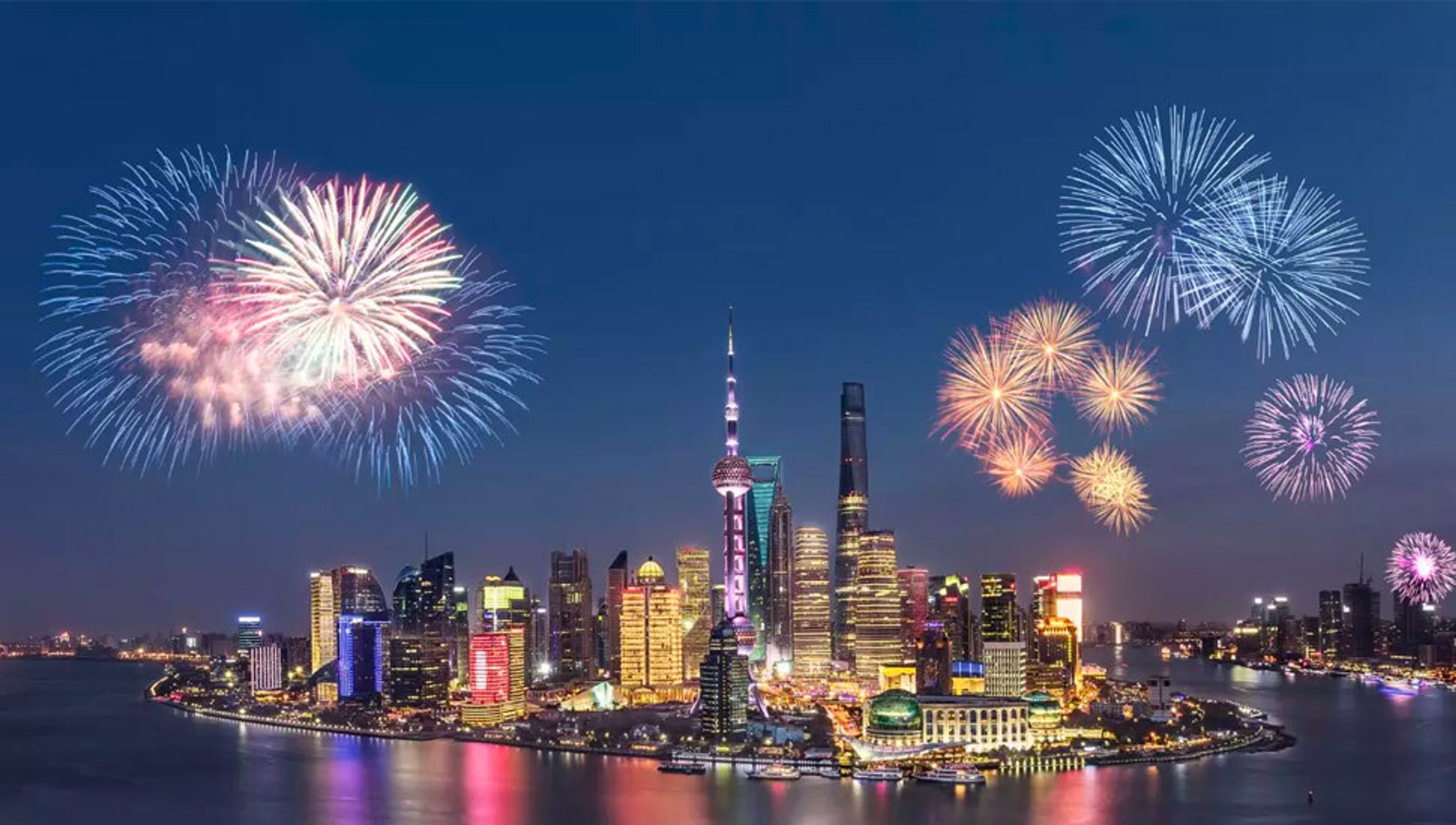 Nighttime view of the Shanghai skyline with colorful fireworks exploding over the river.