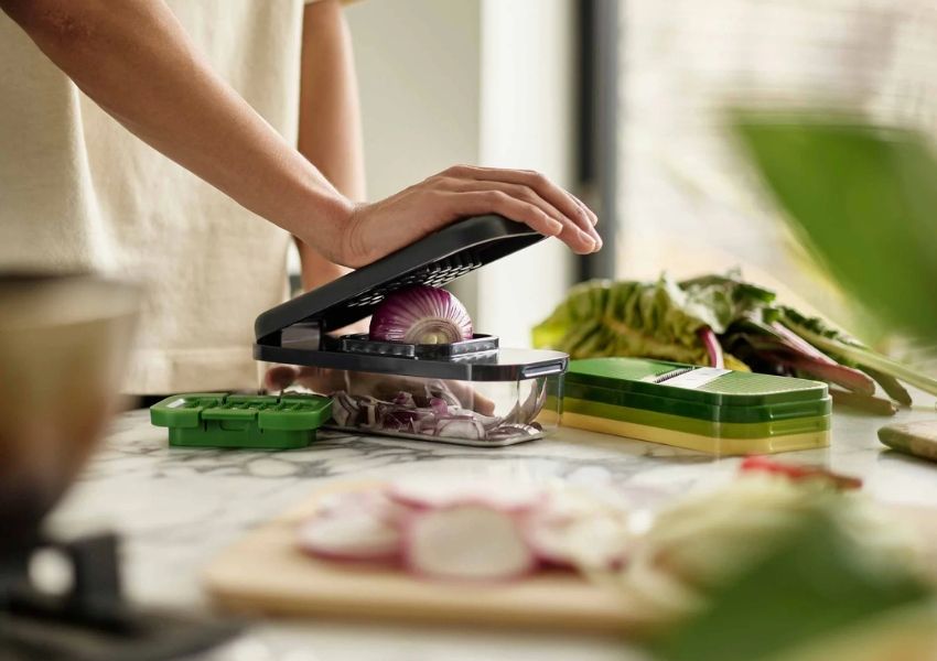 A person uses a black vegetable chopper to dice a red onion on a marble counter.