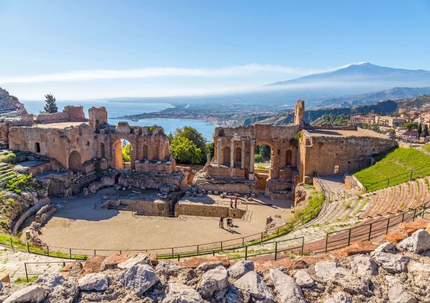 Ancient Greek theater ruins overlooking a coastal town and bay, with Mount Etna in the background under a clear blue sky.