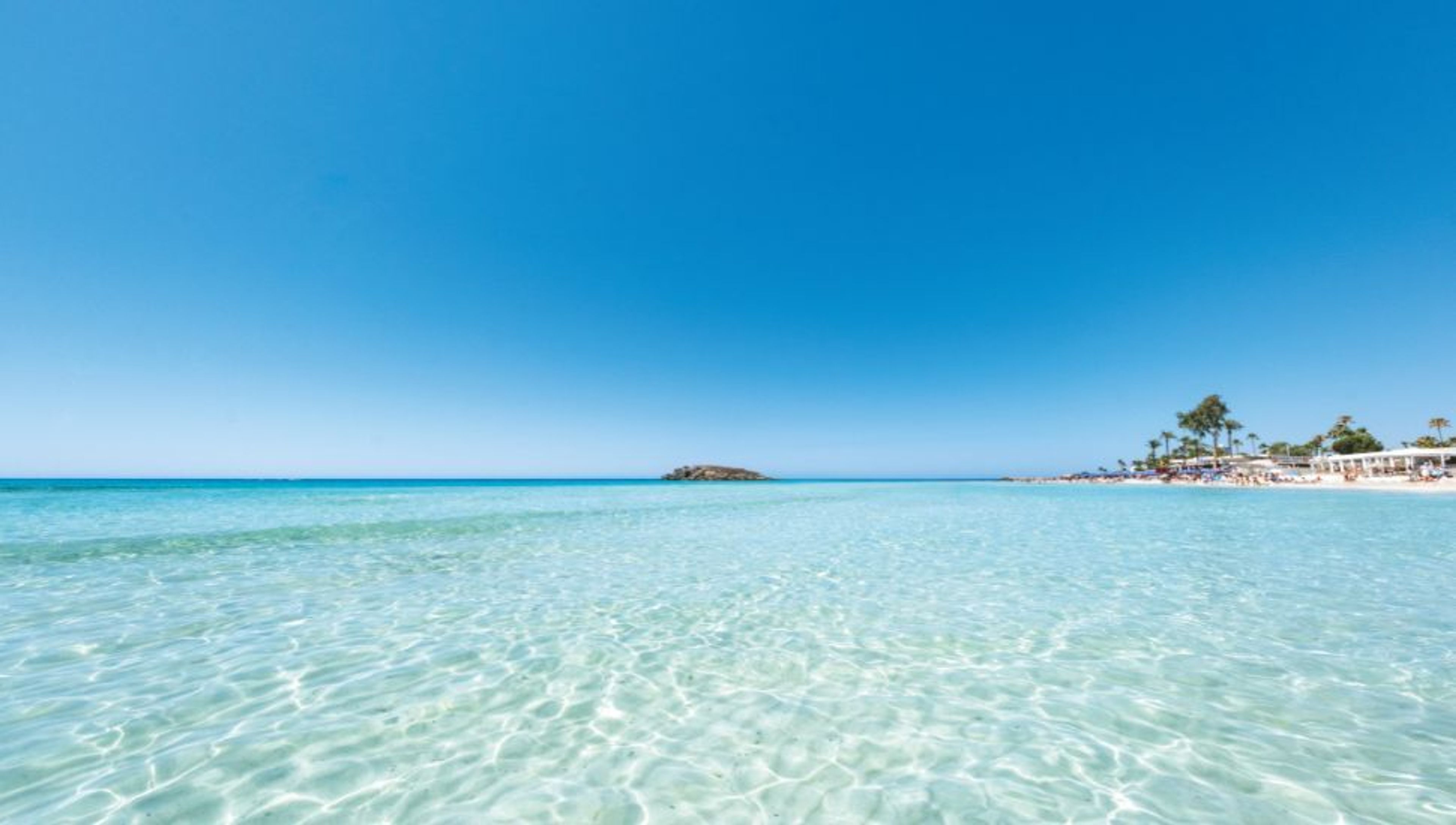 Clear turquoise water under a bright blue sky, with a distant beach and small rocky island.