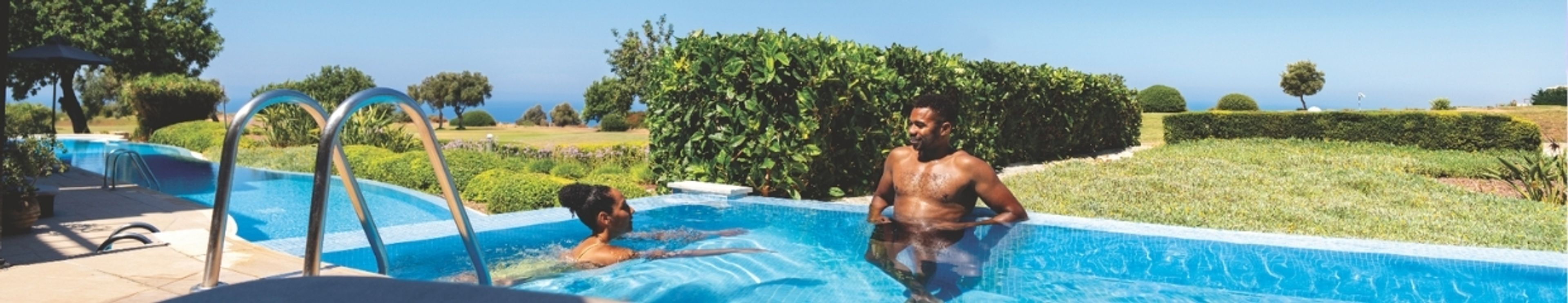 A man and woman relax in an outdoor swimming pool surrounded by lush greenery under a clear sky.