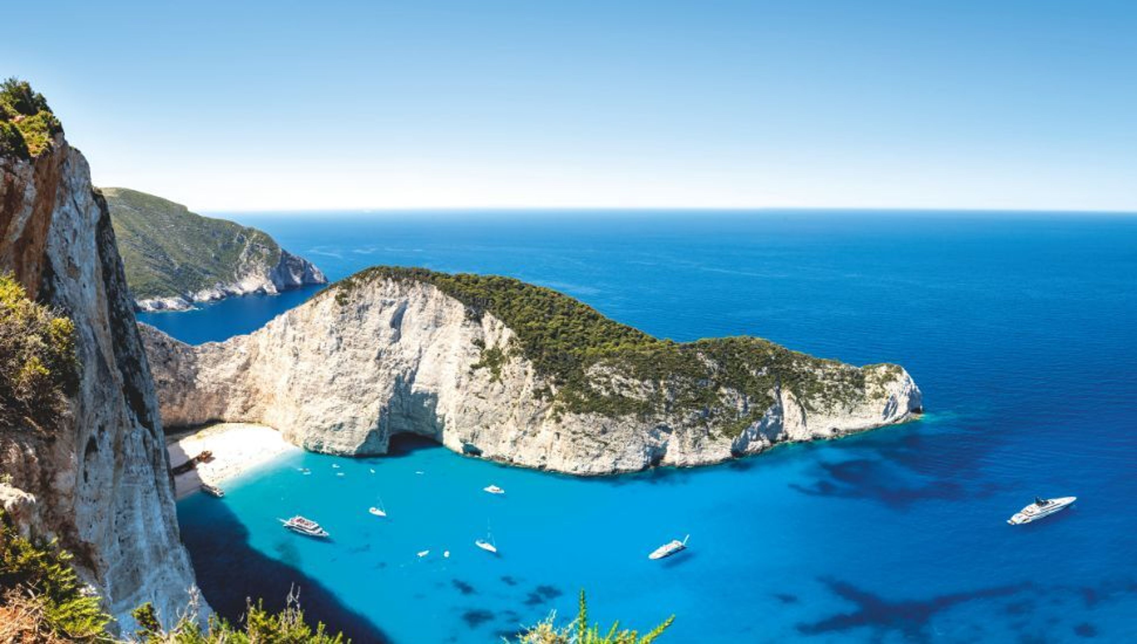 White cliffs frame a sandy beach and turquoise bay dotted with boats under a clear blue sky.