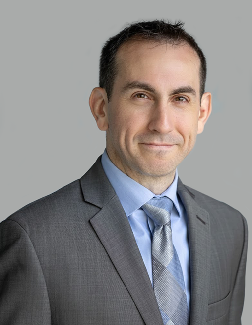 Portrait of a man in a gray suit, light blue shirt, and patterned tie against a gray background.